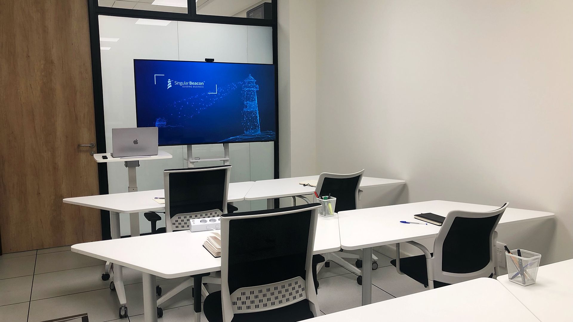 Aula con mesas blancas, sillas y una pantalla grande al fondo